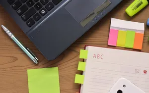 An open address book with alphabet tabs lying on a wooden desk beside a laptop, a pen, sticky notes and a highlighter.