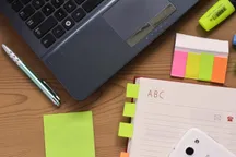 An open address book with alphabet tabs lying on a wooden desk beside a laptop, a pen, sticky notes and a highlighter.