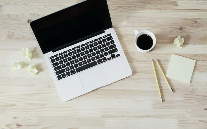 A laptop seen from above on a pale wooden desk, next to a cup of black coffee, two pencils, a pad of sticky notes and a few crumpled paper notes.