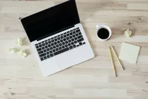 A laptop seen from above on a pale wooden desk, next to a cup of black coffee, two pencils, a pad of sticky notes and a few crumpled paper notes.